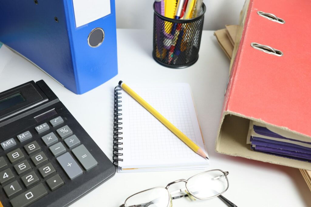 Calculator, blue and coral binders, a notebook with a yellow pencil, and glasses on a white desk.