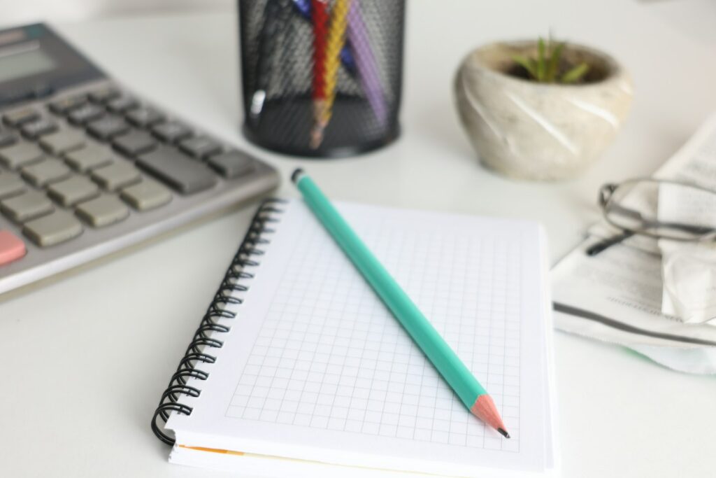 Calculator beside an open notebook with a green pencil, glasses and a small potted plant.